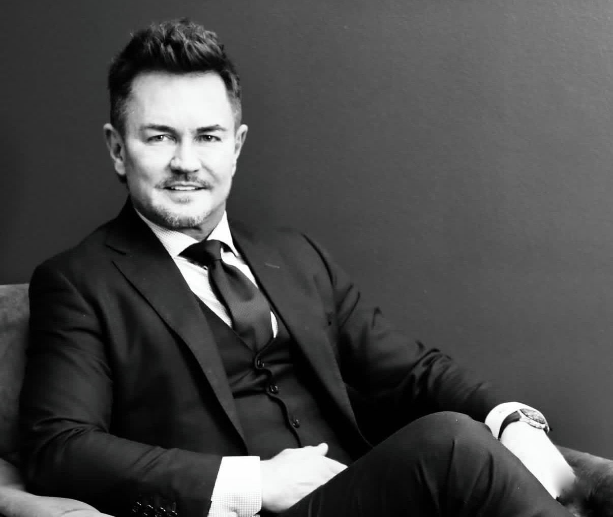 Man in a suit sitting in a chair against a plain background, smiling slightly.
